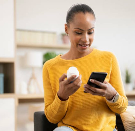 Woman holding medicine searching information online by smartphone sitting on sofa in living room at home.